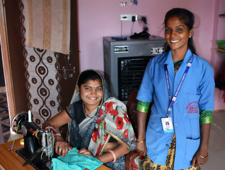 A woman, sitting by her tailoring machine, poses for the camera with a community health worker.