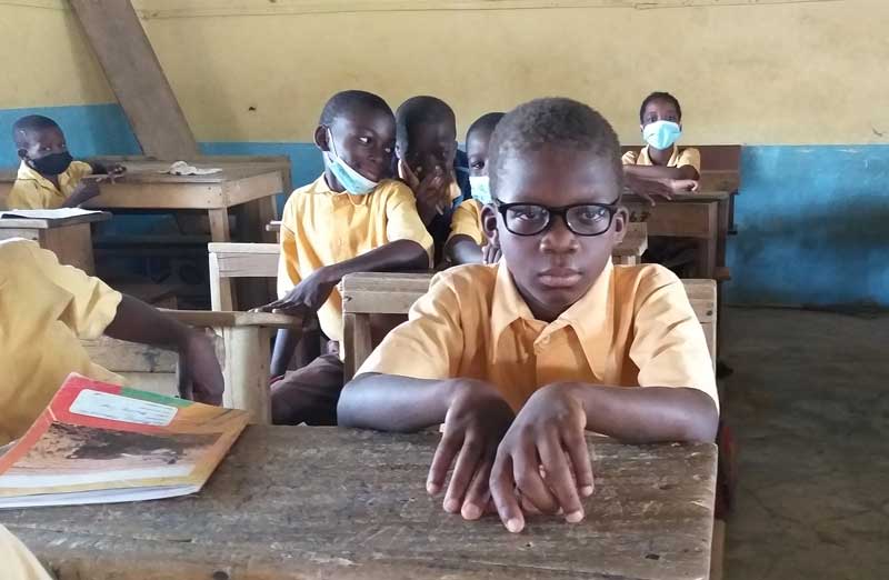 A young boy, wearing eyeglasses, sits in his classroom, surrounded by his peers