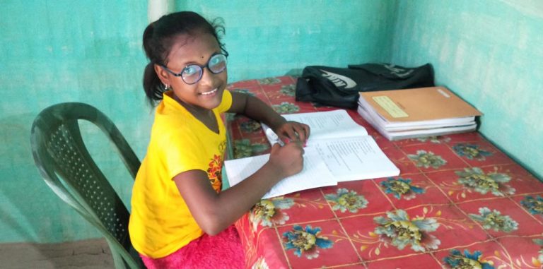 A young girl, wearing eyeglasses and seated at her desk, smiles at the camera as she studies her textbook.