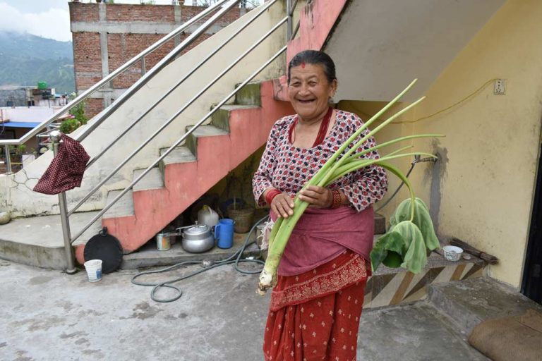A senior woman, holding a vegetable, smiles at the camera in front of her house