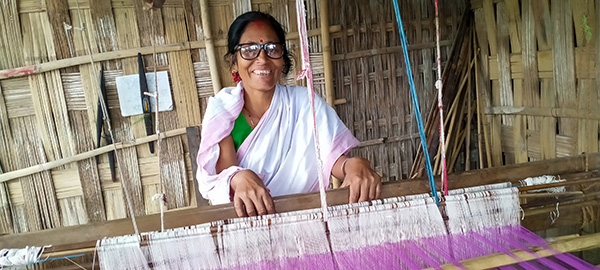 A woman, wearing eyeglasses, poses for the camera as she works on her handloom weaver.