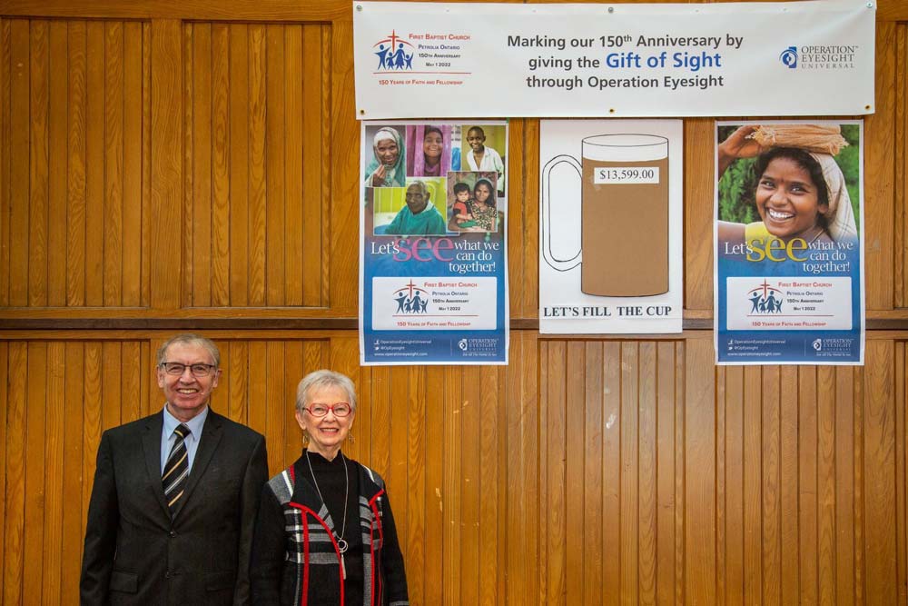 A senior woman and man post for a photo in front of a few posters that show a fundraising campaign for Operation Eyesight and its progress. The text on one of the posters reads "Making our 150th anniversary by giving the gift of sight through Operation Eyesight."