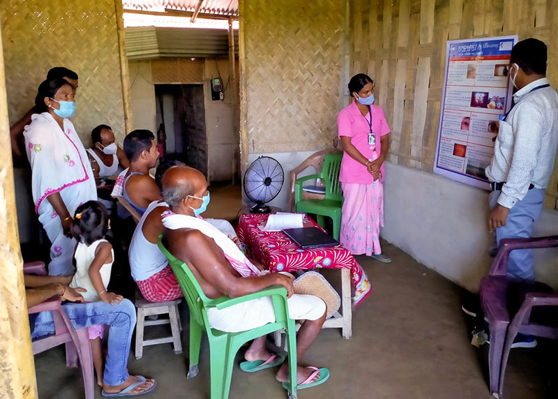 A community health worker educates a group of people on eye health using a poster affixed to a wall