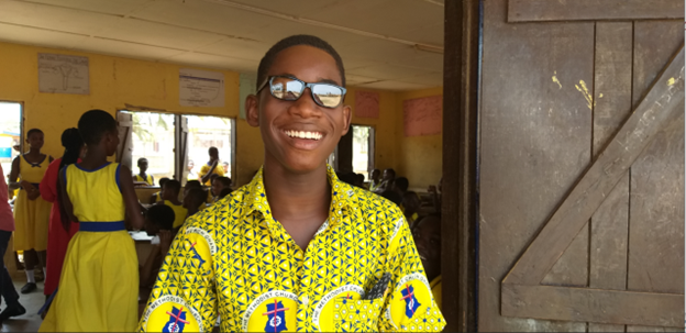 A student, wearing eyeglasses, smiles at the camera. In the background is his classroom and classmates.
