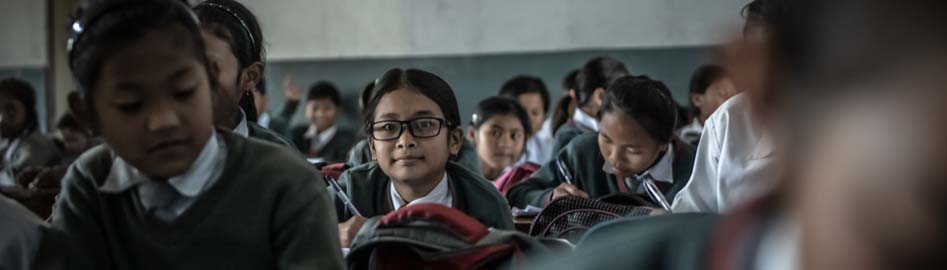 A female student, wearing eyeglasses, sits in her classroom along with her peers and looks at the camera