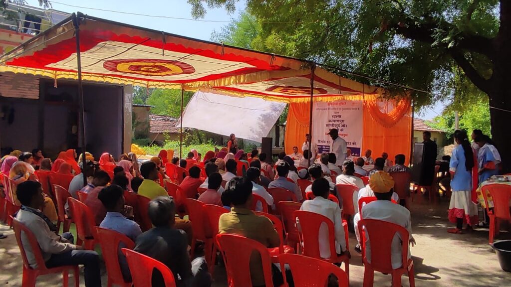 Community members from Madhya Pradesh gather in red chairs facing a celebration banner. At the right of the frame stand two community health workers, donning blue shirts.