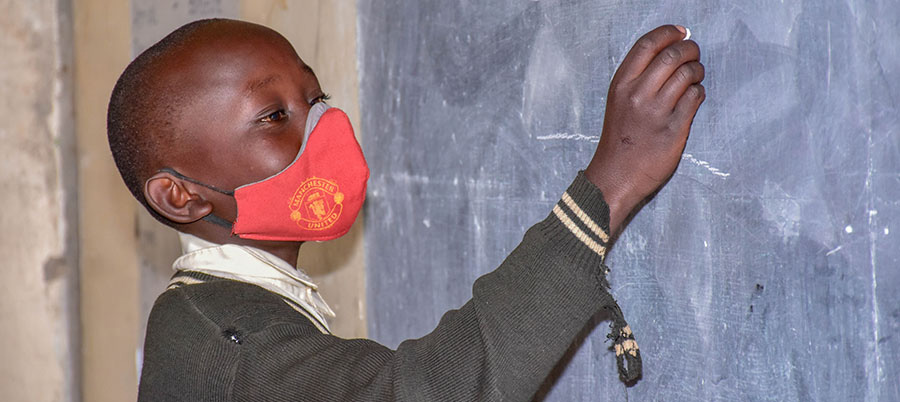 a student, wearing a mask, writes on black board with chalk.