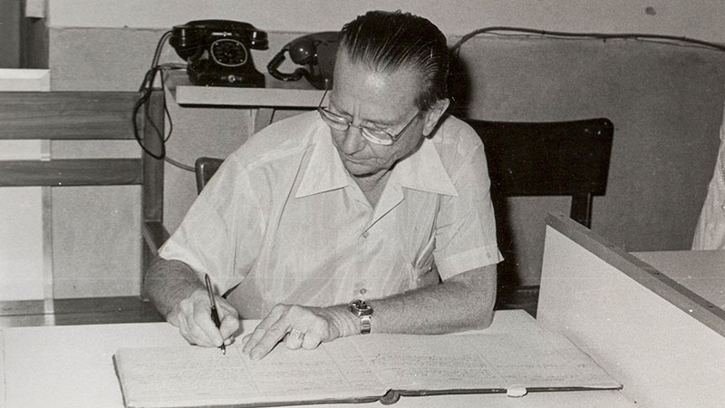 A black and white historical photo shows Art Jenkyns busily working at his office desk.