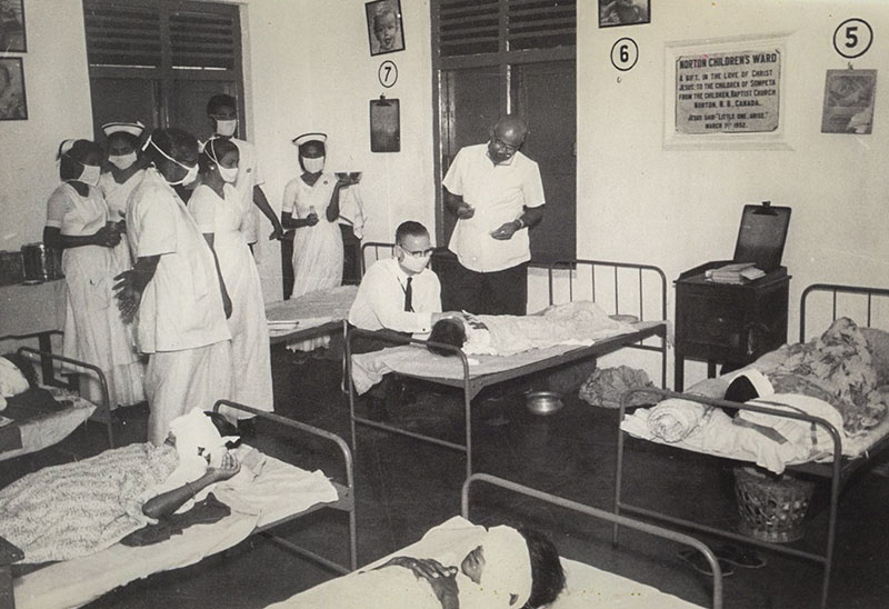 A historical black and white photo shows a man at the bedside of a child patient in a hospital room, surrounded by medical staff.  