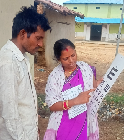 Female health worker wearing a purple sari stands beside a male patient wearing a white shirt, as she conducts a vision test.