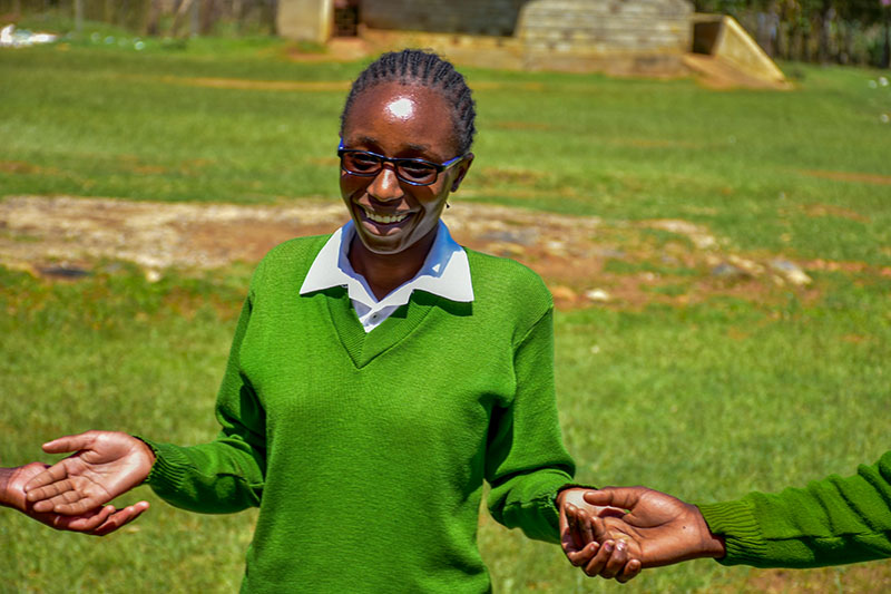 A teenage girl wearing a green school uniform sweater and blue eyeglasses stands hand-in-hand with friends, smiling. 