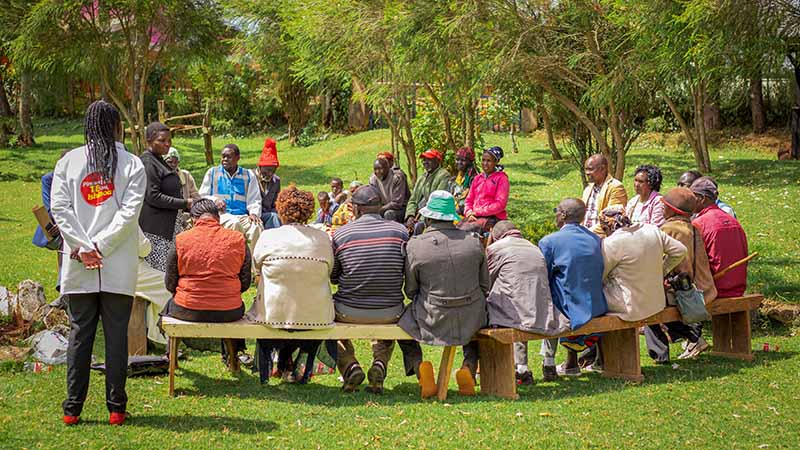 A group of people sit on benches and stand, listening and talking, in a park-like setting, as part of a community meeting in Elgeyo Marakwet, Kenya.