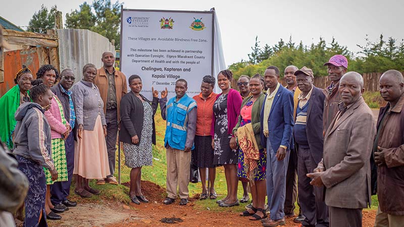 A group of 19 men and women pose in front of a large poster announcing an Avoidable Blindness-Free Zone in a rural setting in Elgeyo Marakwet, Kenya. 