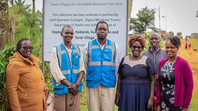 Three women and three men pose in front of a large poster announcing an Avoidable Blindness-Free Zone in a rural setting in Elgeyo Marakwet, Kenya.