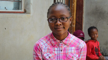 A teenage girl smiles at the camera, sitting outside a building. She wears a pink and white plaid blouse and tortoiseshell eyeglasses.