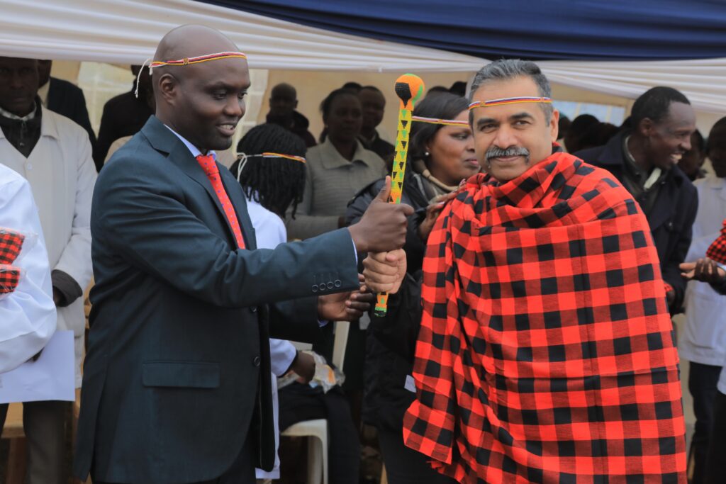 A man in a suit hads a brightly decorated staff to a man with a moustache, wearing a traditional red and black Kenyans cloak. 