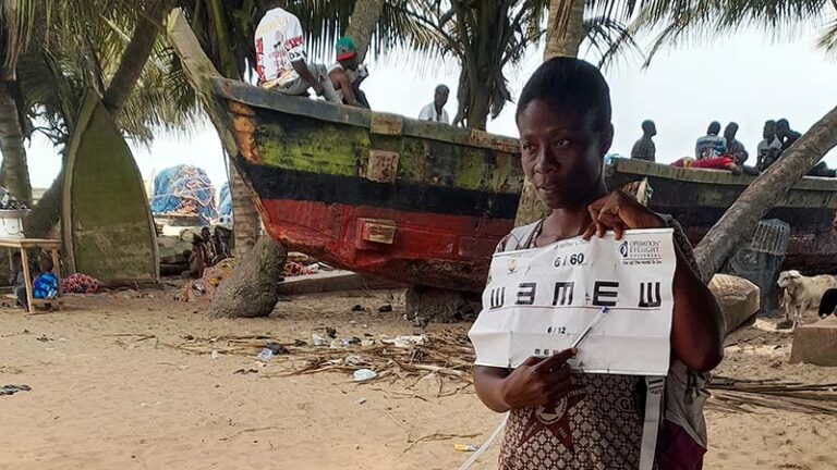 A woman holds up an eye chart to people to people not seen in the photo. Behind her, you can see sandy terrain, palm trees and a fishing boat with several men on board.