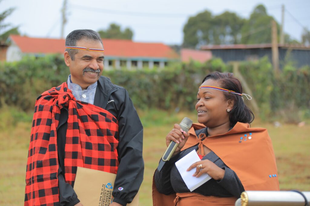 A gentleman with a moustache, wearing a traditional red and black plaid cloak, smiles as he looks at a woman, holding a microphone.