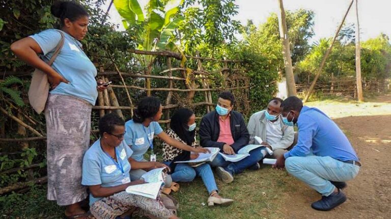: A group of community health workers wearing matching blue t-shirts meet with Yashwant Sinha and some program managers in the shade beneath a fence in a rural setting in Bahir Dar town, Ethiopia.