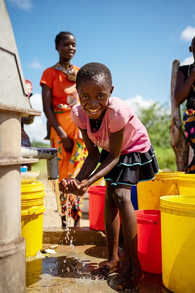 Smiling young girl washes hands at water pump, woman smiles in the background.