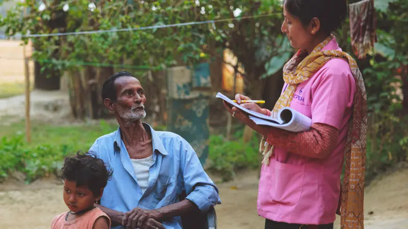Female health worker wearing pink, fills out a form while speaking with an elderly man sitting down with a young boy.