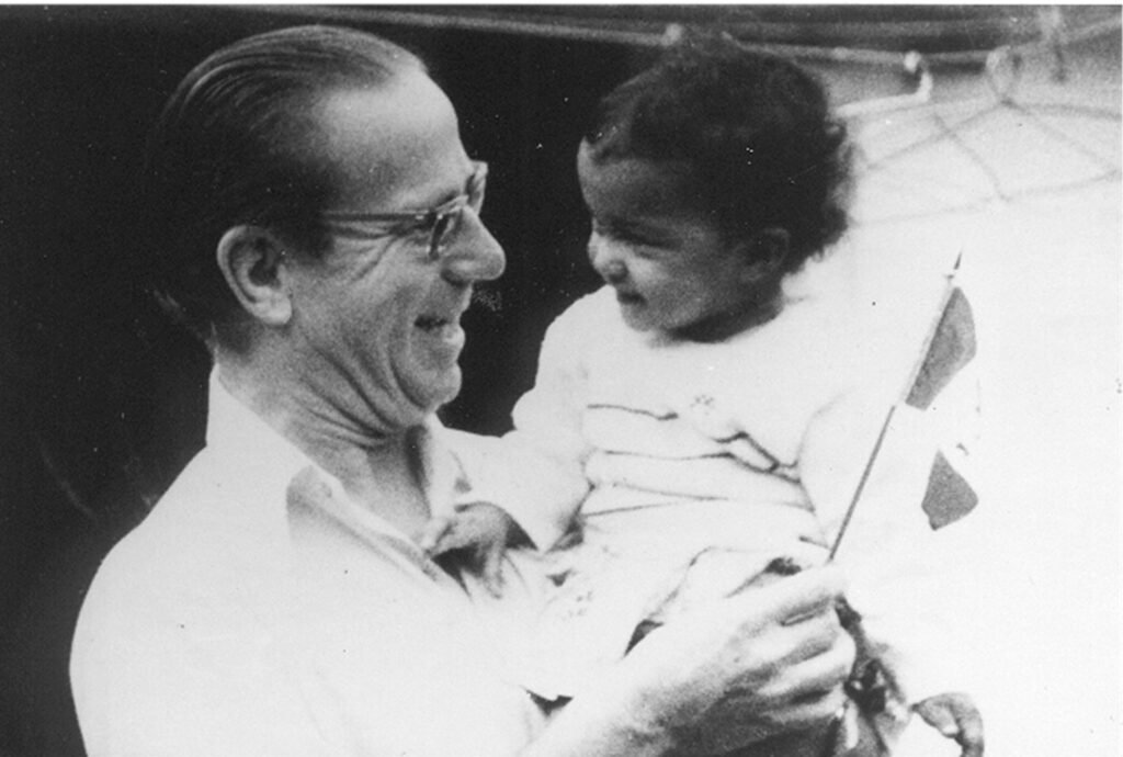 Black and white image of a man with glasses holding a young child in his arms. They are smiling at each other. 