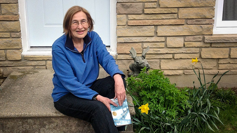 Woman sits on the front steps of her home, holding a mystery novel.