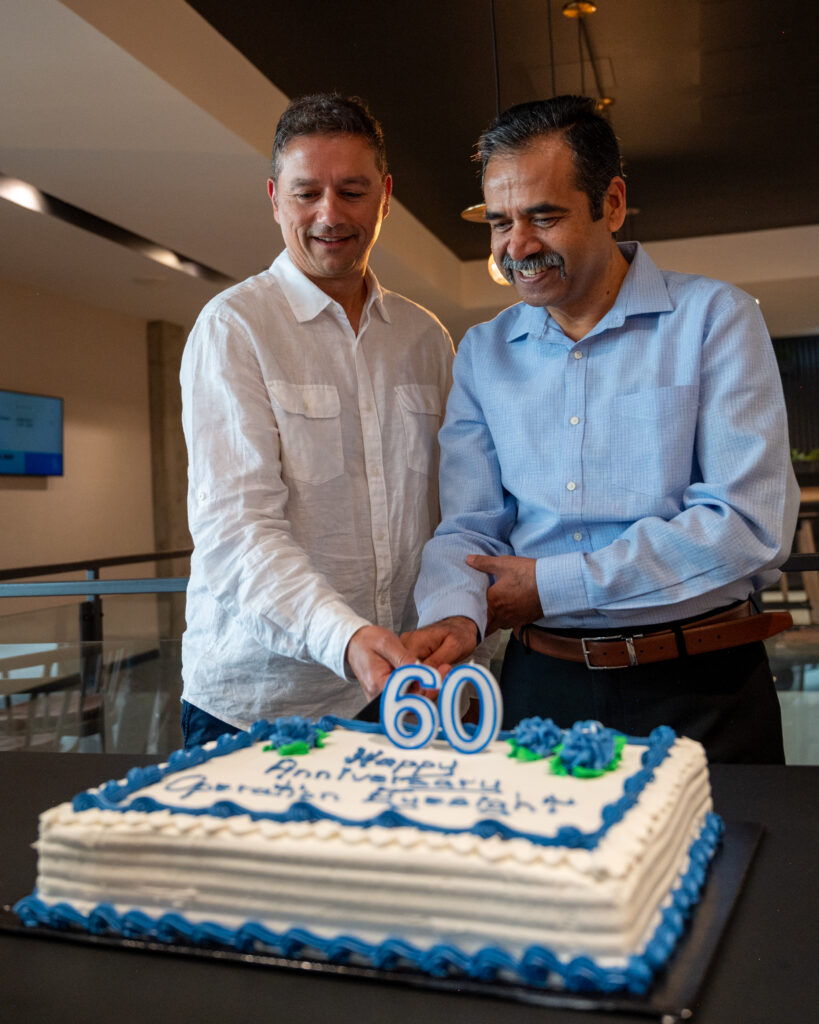 Two men cut a cake with candles shaped like the number 60.