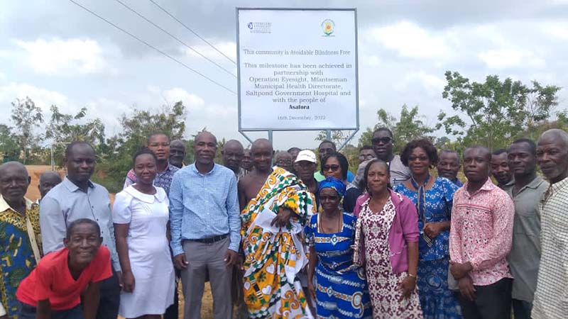 Community members and dignitaries stand in front of an Avoidable Blindness-Free village billboard.