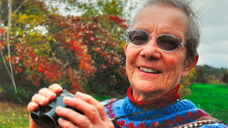 Woman wearing sunglasses holds up binoculars, with tress in the background.
