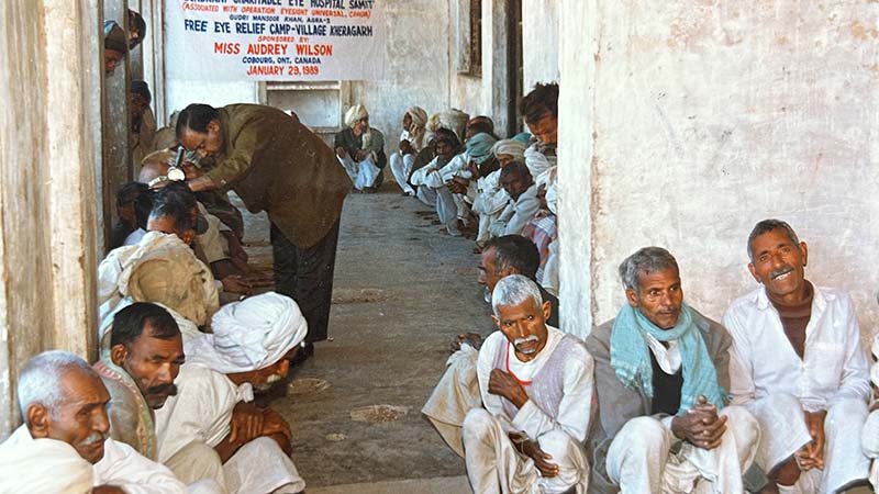 Male patients sitting along a wall, with a banner advertising an eye camp in the background.