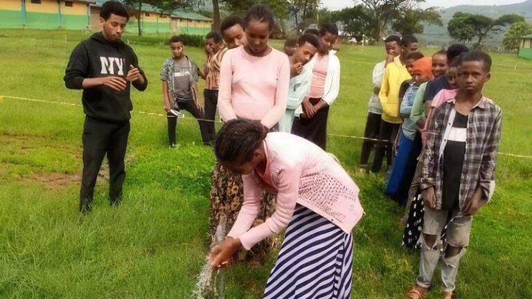 Children line up in a green field in front of a water tap. In the foreground a girl washes her hands.