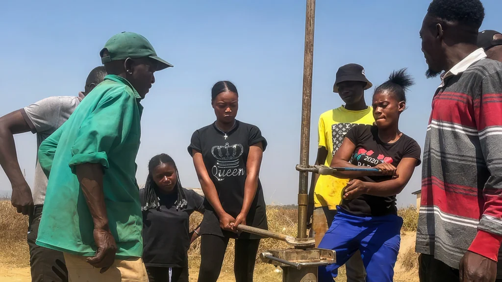 A group of men and women stand around a water borehole pump. Two women appear to be turning a pipe using large wrenches.