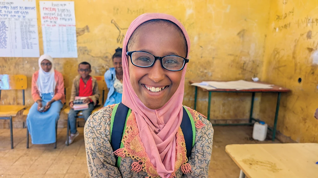 A girl wearing a scarf and black eyeglasses grins at the camera. She is in a classroom. A student and teacher sit in the background.