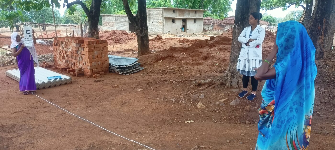 A community health worker checks the eyesight of a community member using an eye chart.|Female health worker wearing a purple sari stands beside a male patient wearing a white shirt