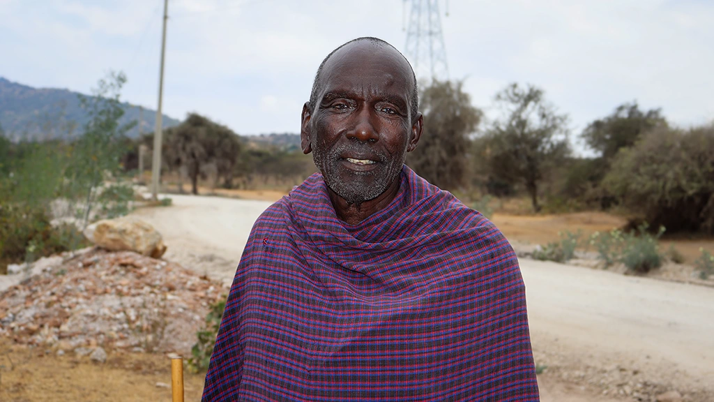 A man wearing a plaid wrap poses for a photo outside. You can see a dirt road