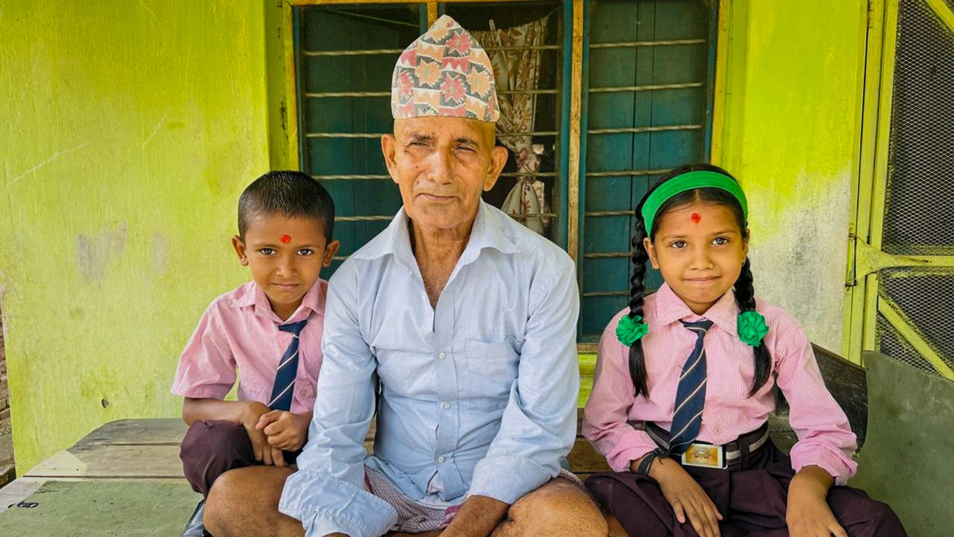 A man wearing a Nepali topi hat poses with two young children in school uniforms.|