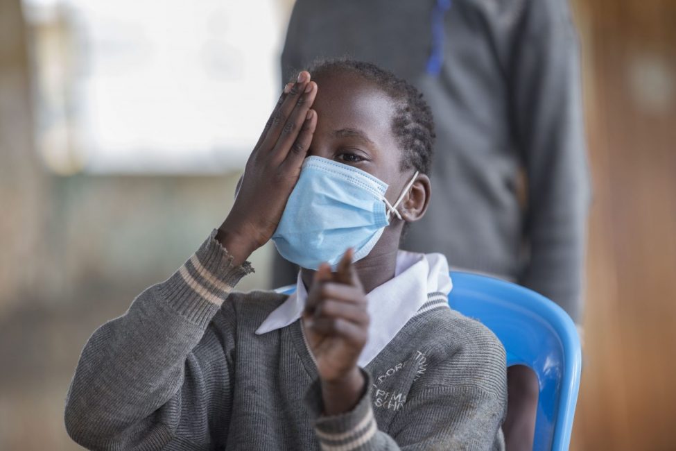 Child covers her right eye with her right hand. She points towards the camera with her left hand as she looks through her left eye.|A student