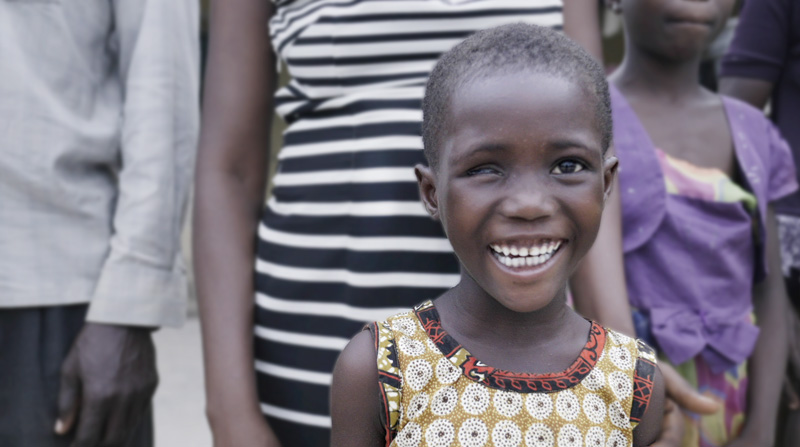 A photo of a child with visual impairment smiles at the camera with other members of her community standing behind her.|A child with visual impairment poses in front of a hospital for a photo with her father and Operation Eyesight's Country Director for Ghana.