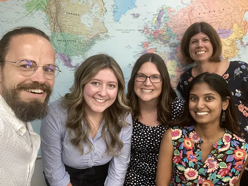 Photo of a group of people as they pose for the camera in front of a world map.|A screenshot of a group of people from an online video meeting.